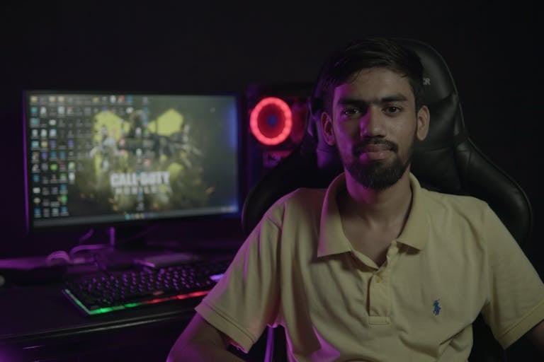 A bearded man sitting at a computer setup with a gaming PC and chair, creating a moody ambiance.