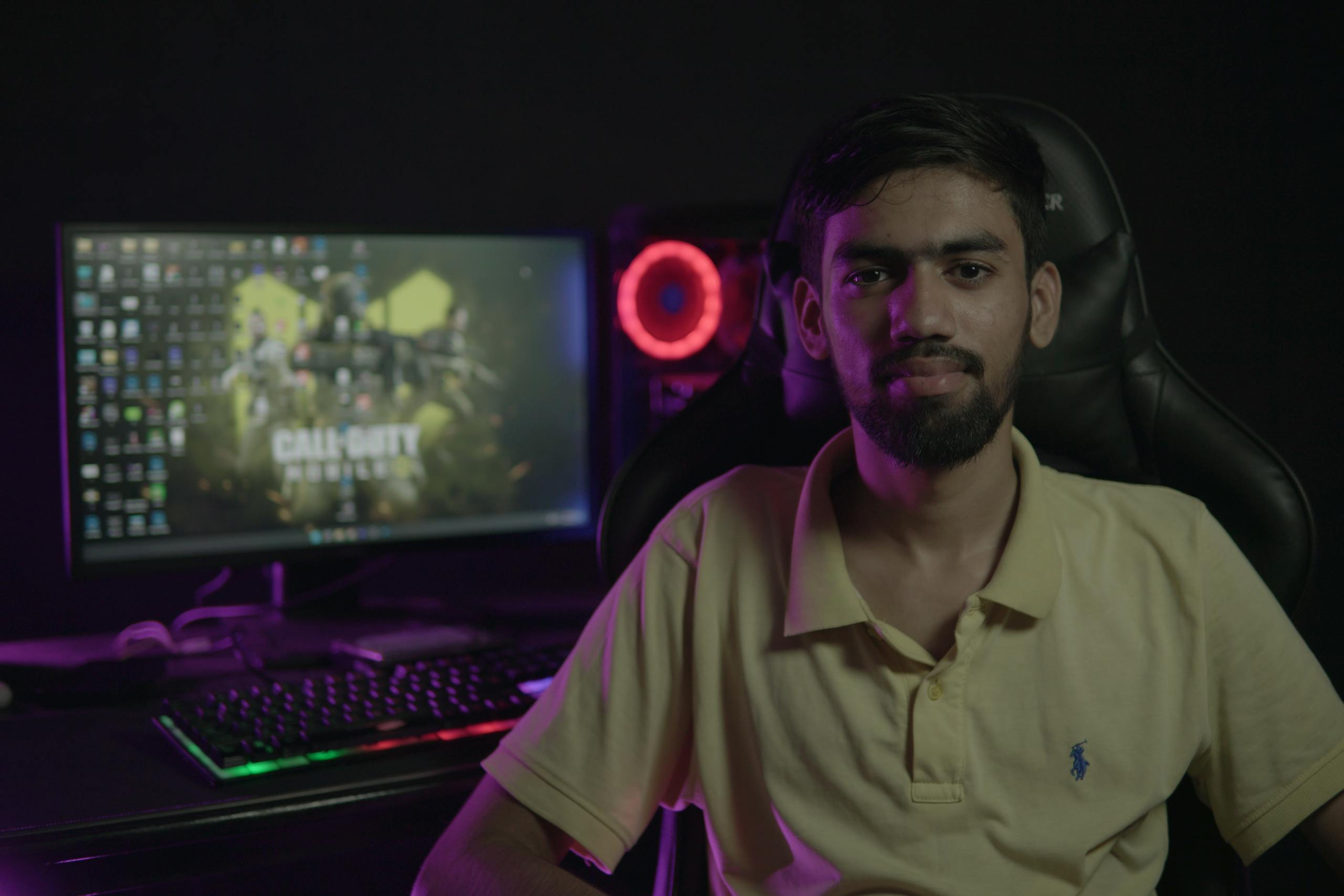 A bearded man sitting at a computer setup with a gaming PC and chair, creating a moody ambiance.
